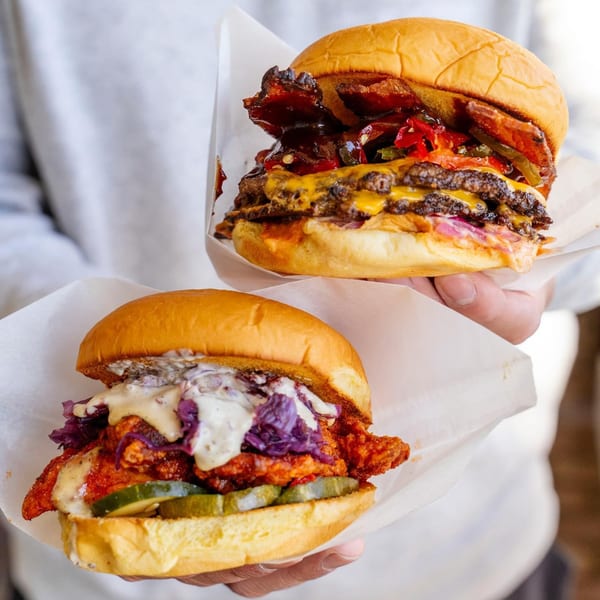 Two hands hold a Konjoe Burger double cheeseburger with bacon and a spicy chicken sandwich.