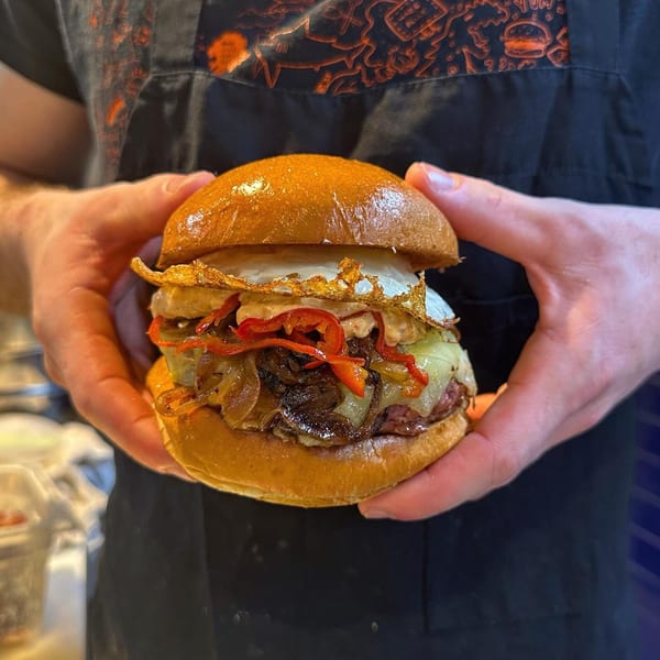 Hands holding a Konjoe Burger with fried egg, caramelized onions, roasted red peppers, and cheese.