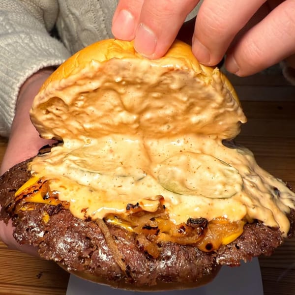Hands holding a Konjoe Burger with a thick patty, melted cheese, caramelized onions, and creamy sauce.