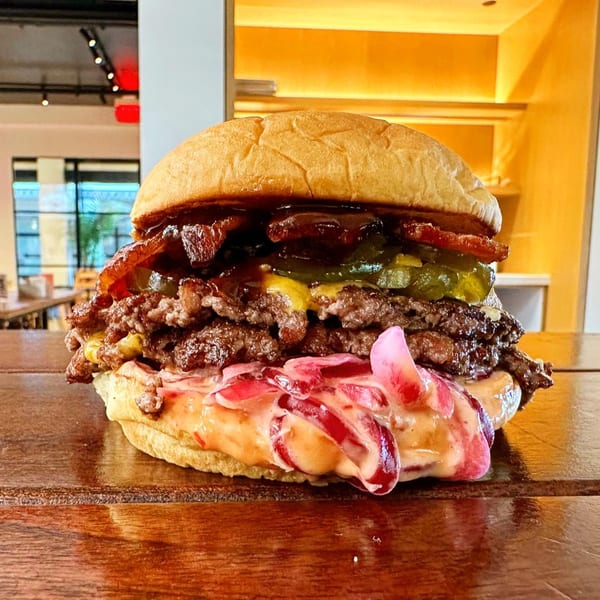 A Konjoe Burger with double patties, bacon, pickles, and pickled onions on a wooden table.