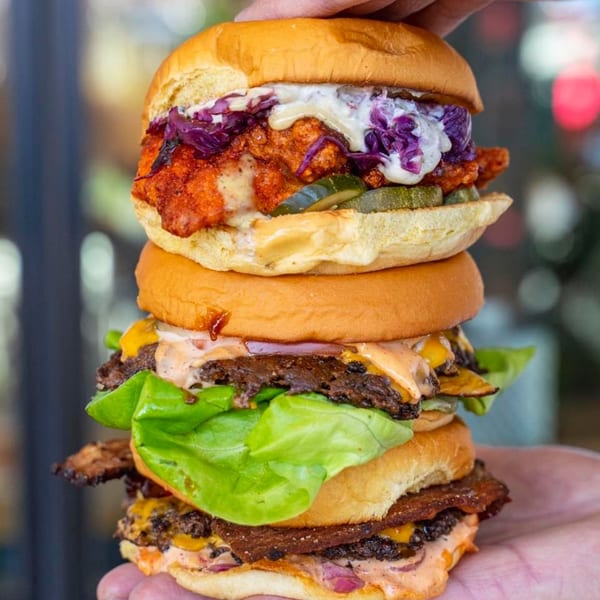A hand holds a towering stack of three Konjoe burgers, each with various toppings.